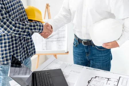 What hidden costs should I expect from a Tampa home remodel? Two construction professionals shake hands over architectural blueprints and a laptop at a project meeting