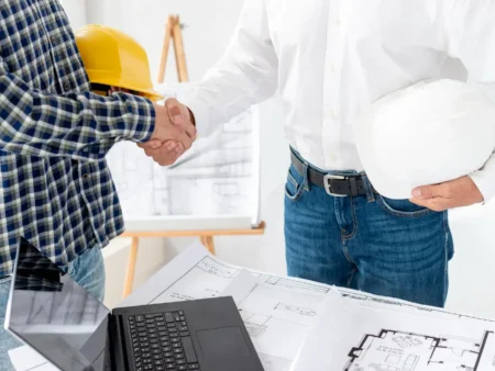 Two construction professionals shake hands over architectural blueprints and a laptop at a project meeting